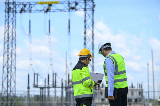 Electrical engineer and technician discuss power grid equipment at outdoor substation, wearing safety gear while reviewing laptop data under bright blue sky in professional setting