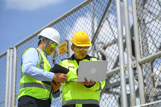 Electrical engineer and construction worker review laptop data beside secure chain link fence during industrial site inspection in bright daylight