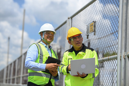 Electrical engineer and construction worker inspecting laptop near industrial fence under blue sky