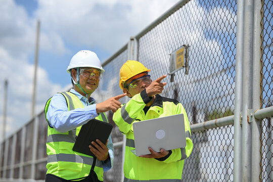 Electrical engineer and technician discussing industrial safety near secure facility fencing under bright cloudy sky