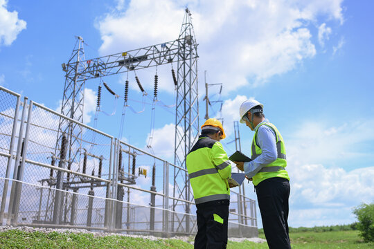 Electrical engineer inspect power substation equipment beside safety fence under blue sky, supporting teamwork and careful industrial maintenance in outdoor energy facility
