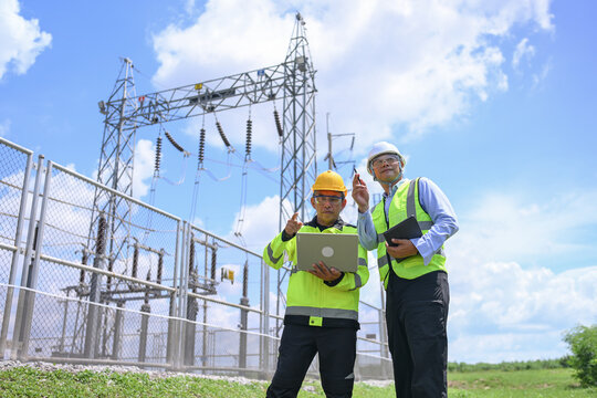 Electrical engineer inspecting substation equipment with coworker, laptop, safety helmet, and reflective vest under bright blue sky, conveying teamwork and careful professional monitoring