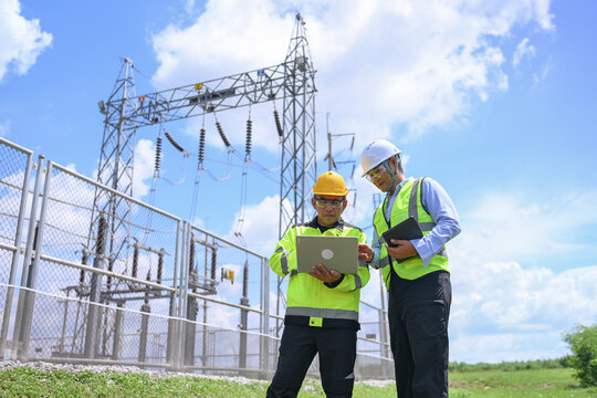 Electrical engineer inspecting substation equipment with tablet beside power transmission tower under bright blue sky