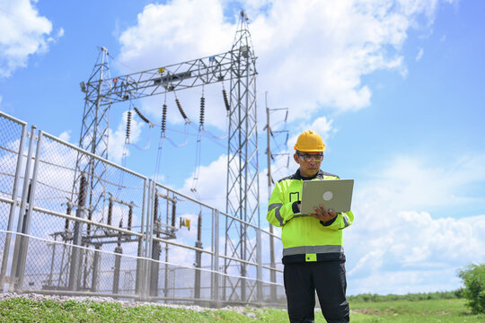Electrical engineer inspecting power substation equipment outdoors under bright blue sky, using laptop for safe infrastructure maintenance and energy system analysis