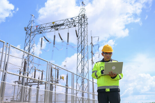 Electrical engineer inspecting substation equipment with laptop beside fenced power transmission facility under bright blue sky