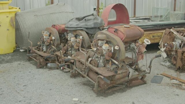 A row of rusted engine assemblies, fuel tanks and other mechanical scrap is piled outdoors at a salvage yard beside rolls of wire mesh and plastic bins.