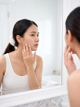 Young woman in white tank top touching acne on cheek while looking in bathroom mirror, skin concern and self-image