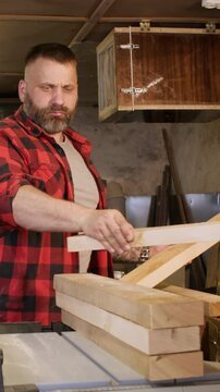 Vertical video. Joinery process. Stacking wood. Bearded carpenter placing freshly cut blocks on table saw in garage workshop with tools set on wall.