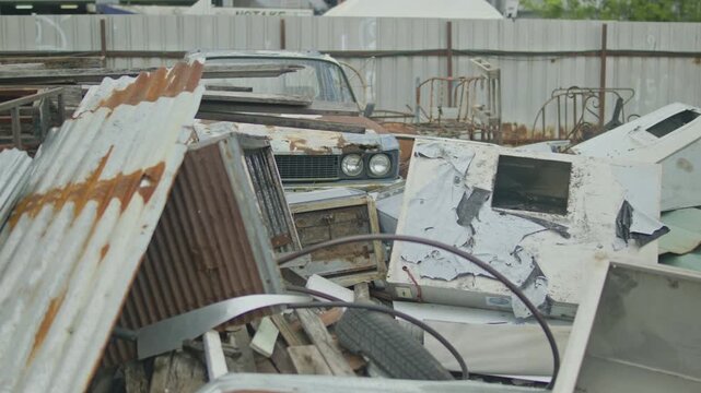 A camera pan across a scrapyard reveals rusted corrugated metal sheets, discarded appliances, tires and the front of an old abandoned car amid piled debris.