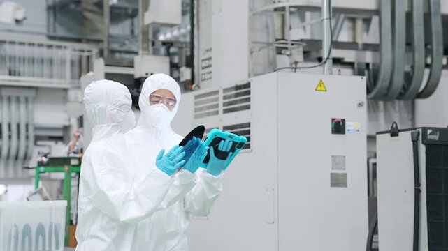 半導体工場でタブレットを持ってシリコンウエハーを点検する防護服を着た男女作業員