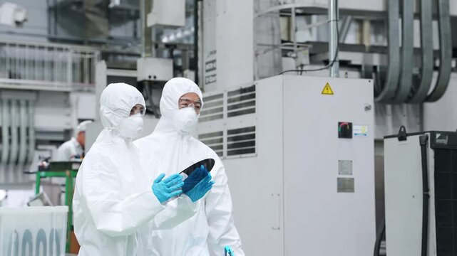 Male and female workers in protective suits inspect silicon wafers with tablets in a semiconductor factory.