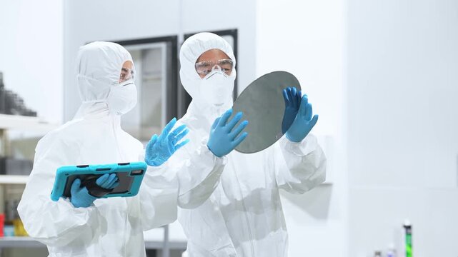 Male and female workers in protective suits inspect silicon wafers with tablets in a semiconductor factory.