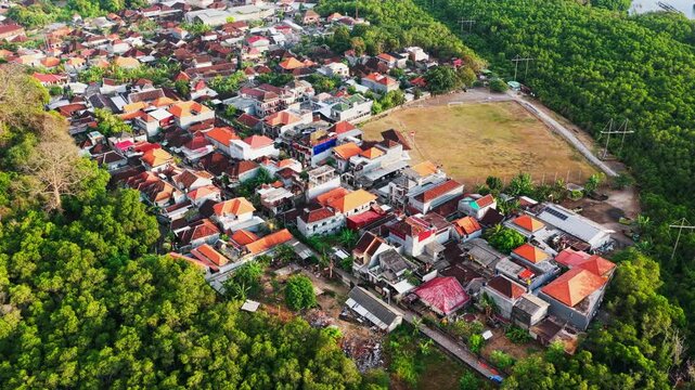 Dense residential neighborhood with red tile roof houses bordered by a lush green tropical mangrove forest and open grass soccer field overhead drone footage