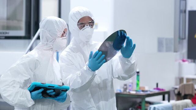 Male and female workers in protective suits inspect silicon wafers with tablets in a semiconductor factory.