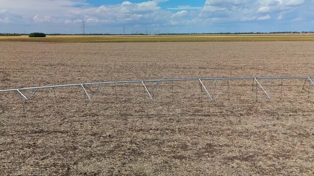 A drone tracks along a center-pivot irrigation system across a dry harvested field near Debrecen, Hungary, with power lines and cropland beneath a partly cloudy summer sky.