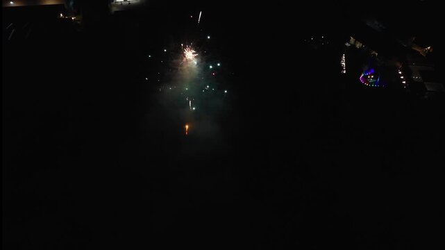 Drone view of one firework shell bursting white and orange at night, leaving red smoke over lit buildings below