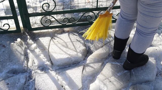 Woman cleaning the path from snow with broom. Worker clearing pathway from frost ice with broom, sweeping. Suburban backyard in winter season. Cold weather