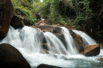 A vibrant, multi-tiered waterfall cascading over a series of dark, jagged rocks within a lush, tropical jungle environment. © k33pcalmncapture