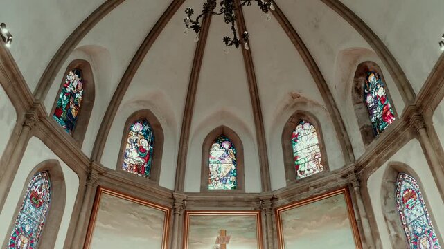 Apse of a church with ribbed vaulting, stained glass windows and painted panels framing the altar crucifix below.