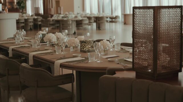 Reception hall with long tables laid for a wedding dinner, white hydrangeas, LED cubes, rattan lanterns and upholstered chairs.