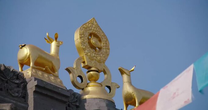 Golden Dharma Wheel flanked by two deer statues atop a temple roof with prayer flags waving in wind against clear blue sky