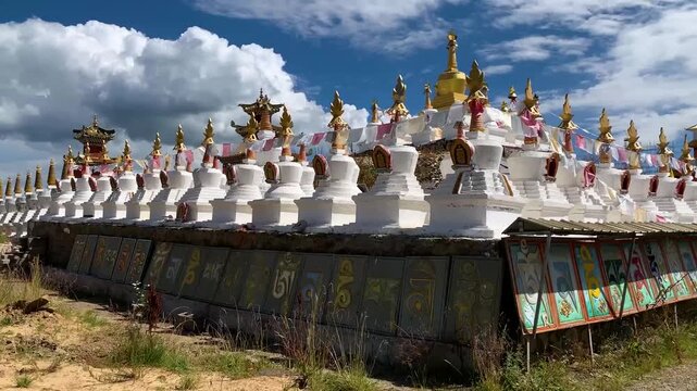 White Tibetan stupas with golden spires and prayer flags lined along a stone wall inscribed with Sanskrit mantras under a bright blue sky with cumulus clouds