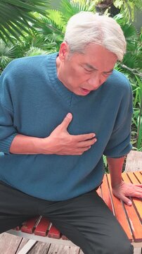 60s senior southeast asian man with white hair suffering from GERD and acid reflux at home, holding his chest with a painful expression on bench in outdoor park, vertical video