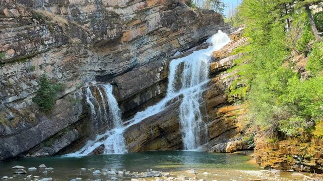 Cameron Falls cascading over layered rock cliffs into a green pool at Waterton Lakes National Park in Alberta, a scenic waterfall surrounded by trees in summer.