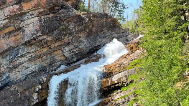 Cameron Falls cascading over layered rock cliffs into a green pool at Waterton Lakes National Park in Alberta, a scenic waterfall surrounded by trees in summer.