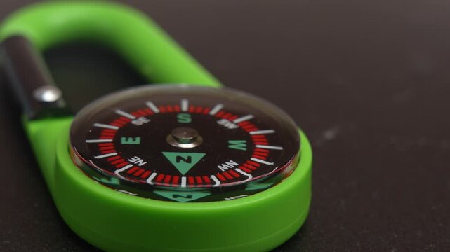 Close-up of a small green carabiner compass with red and white markings lying on a dark surface, filmed with slow, smooth camera movement.