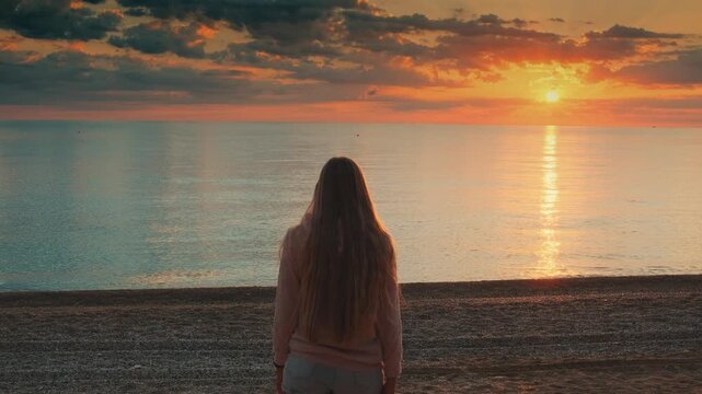 Back view of a woman with long brown hair walking, standing, and posing with outstretched arms on a sandy beach by the calm ocean during daytime.
