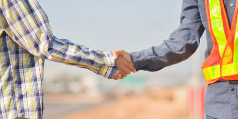 Professional Collaboration in Construction Industry with Two Individuals Shaking Hands in Safety Gear on a Roadside Setting © Suriyo