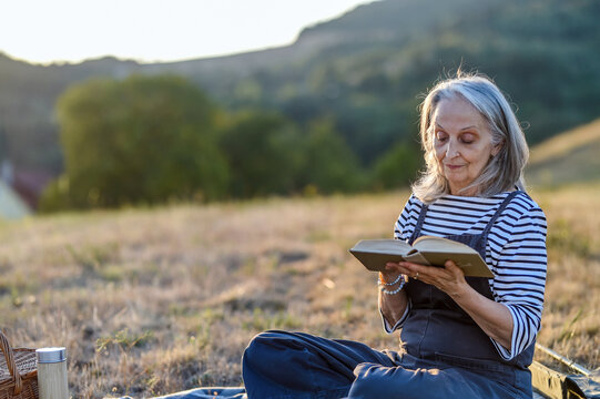 Older woman enjoying peaceful solo picnic and reading in countryside.