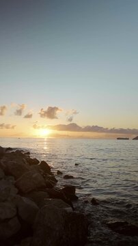 Breathtaking sunset seascape with a vibrant orange-red sky, calm reflective water and a rugged shoreline covered with large coastal stones.