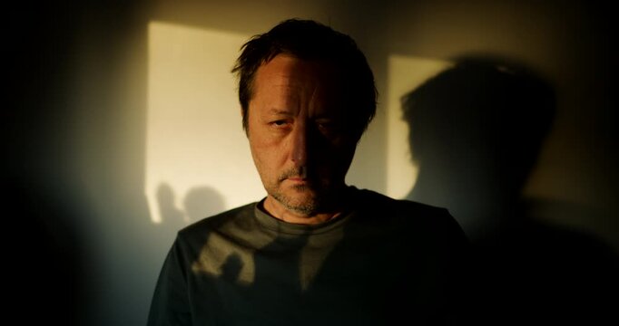 Man standing indoors with dramatic window light casting a strong shadow on the wall behind him, moody portrait. 4K.