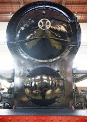 Front view of glossy black steam locomotive with round smokebox door, brass handwheel, and riveted red buffer beam. Preserved heritage railway engine displayed in museum for transport history themes. © Grenar