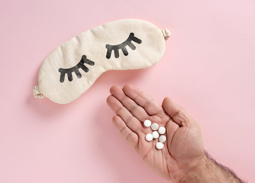 Man's hand holding white sleeping pills near a sleep mask, illustrating insomnia medication, sleep problems and aid concepts