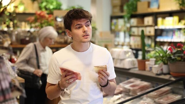 Guy is holding a package of cheese and bacon in his hands in an eco supermarket. European chooses products for breakfast in the organic department of the store . High quality 4k footage