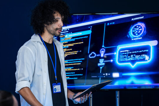 A focused male developer with a curly afro studies an AI assistant interface on the big display alone in a dark blue technology lab at night, an image of human and machine working together