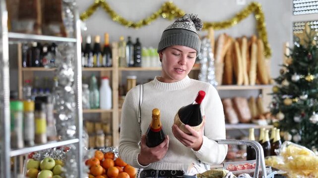 Happy young female shopper in Christmas-themed store with decorated tree and tinsel comparing wine and champagne, choosing drinks for festive gathering 