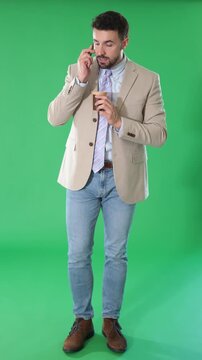 Adult man stands against green screen, holding smartphone to ear and speaking while holding cup of coffee during video recording 
