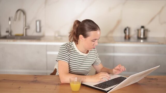 Teenage girl sits against the background of the interior of the kitchen and sadly looks at the screen of the notebook, she is upset that she did not complete the task on time . High quality 4k footage