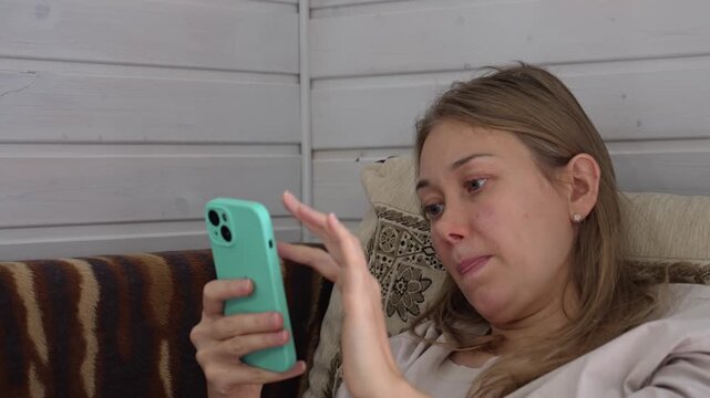 A woman sits comfortably on a sofa while deeply absorbed in her smartphone, mindlessly scrolling through content in a quiet room, reflecting the persistent grip of digital habits on daily life.