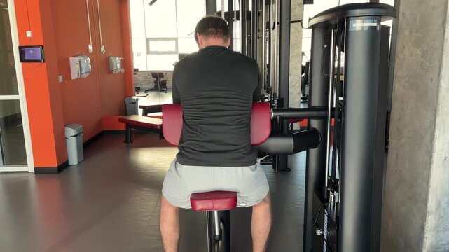 Seen from behind, a man flexes his arms on a preacher curl machine in a gym, biceps workout, resistance training, active lifestyle.