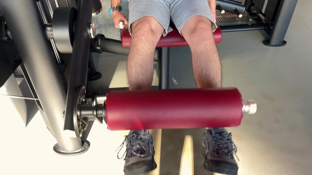 A man lifts a red padded roller with both legs on a seated gym machine, leg extensions, strength training, active lifestyle.