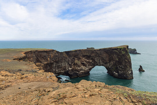 Massive Dyrholaey Arch rises from rugged sea cliffs above the Atlantic Ocean in South Iceland, with offshore rock formations beneath a broad cloudy summer sky and clear horizon.