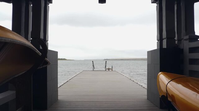 Vertical shot from wooden pier shelter looking out to calm lake. Overturned yellow boats on both sides, boardwalk leading to water, overcast grey sky. Peaceful boating and vacation spot vertical 4k
