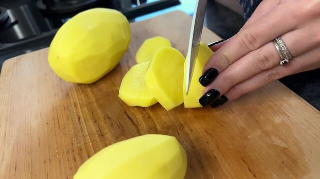 An adult cuts thick slices from a peeled potato in a close view of a kitchen board, knife skills, home cooking, meal preparation.