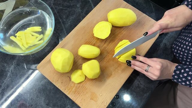 An adult cuts peeled potatoes into thin sticks beside a bowl of water, home cooking, meal preparation, knife skills.