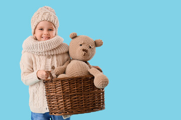 Naklejka na ściany i meble Cute little girl in winter clothes holding basket with toy bear on blue background